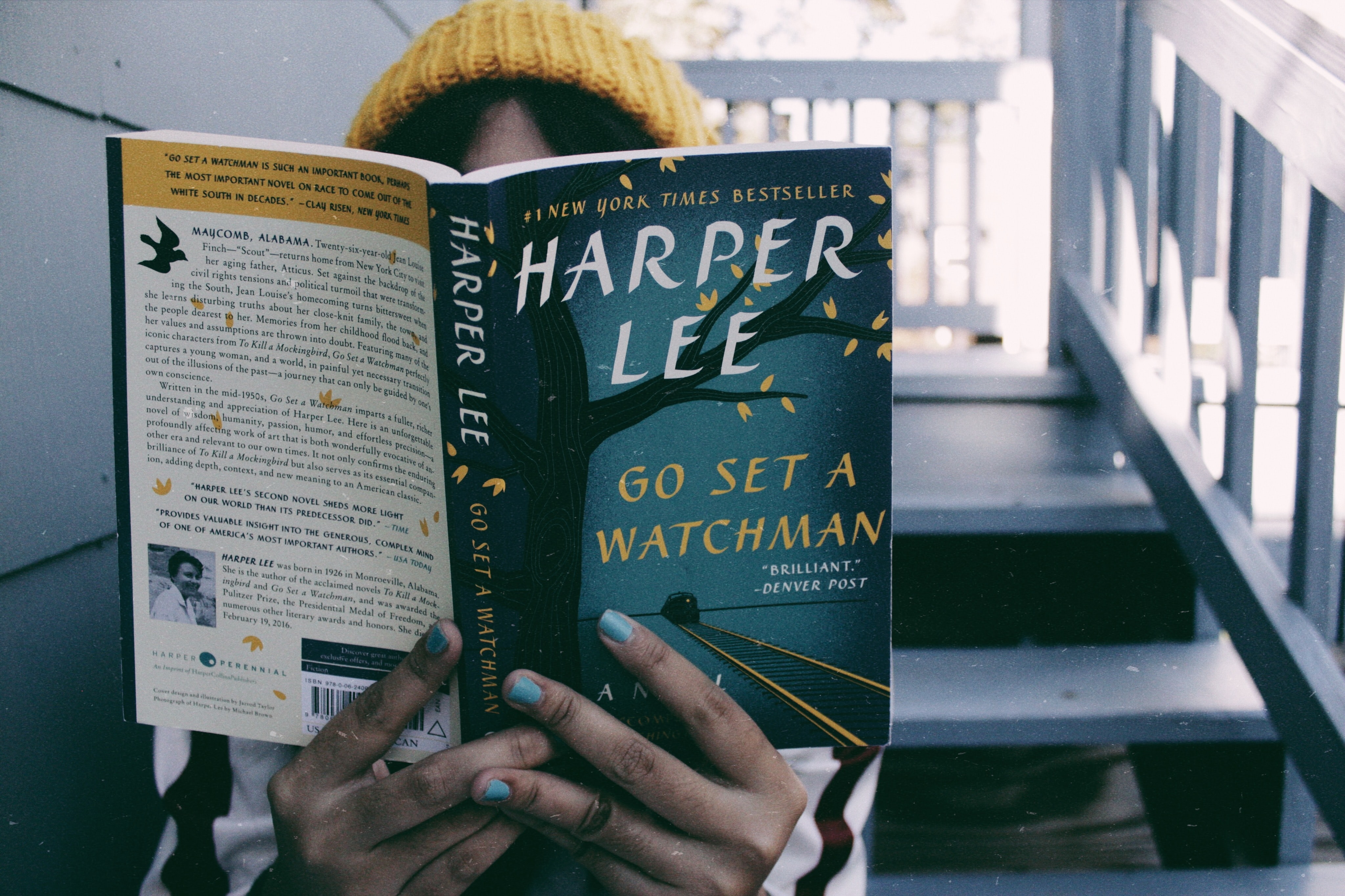 A girl in a yellow hat holds a copy of go set a watchman by harper lee - it is covering her face